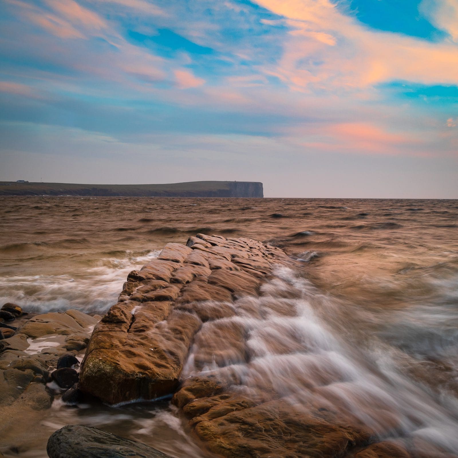 Rocky shoreline extending to a distant cliff; waves swirl on brown sea with pastel sky.